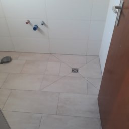 Tomasz - Łazienka w trakcie wykańczania: jasne płytki podłogowe i ścienne, widoczne rury instalacji wodnej i kanalizacyjnej, odpływ liniowy w podłodze, fragment brązowych drzwi.