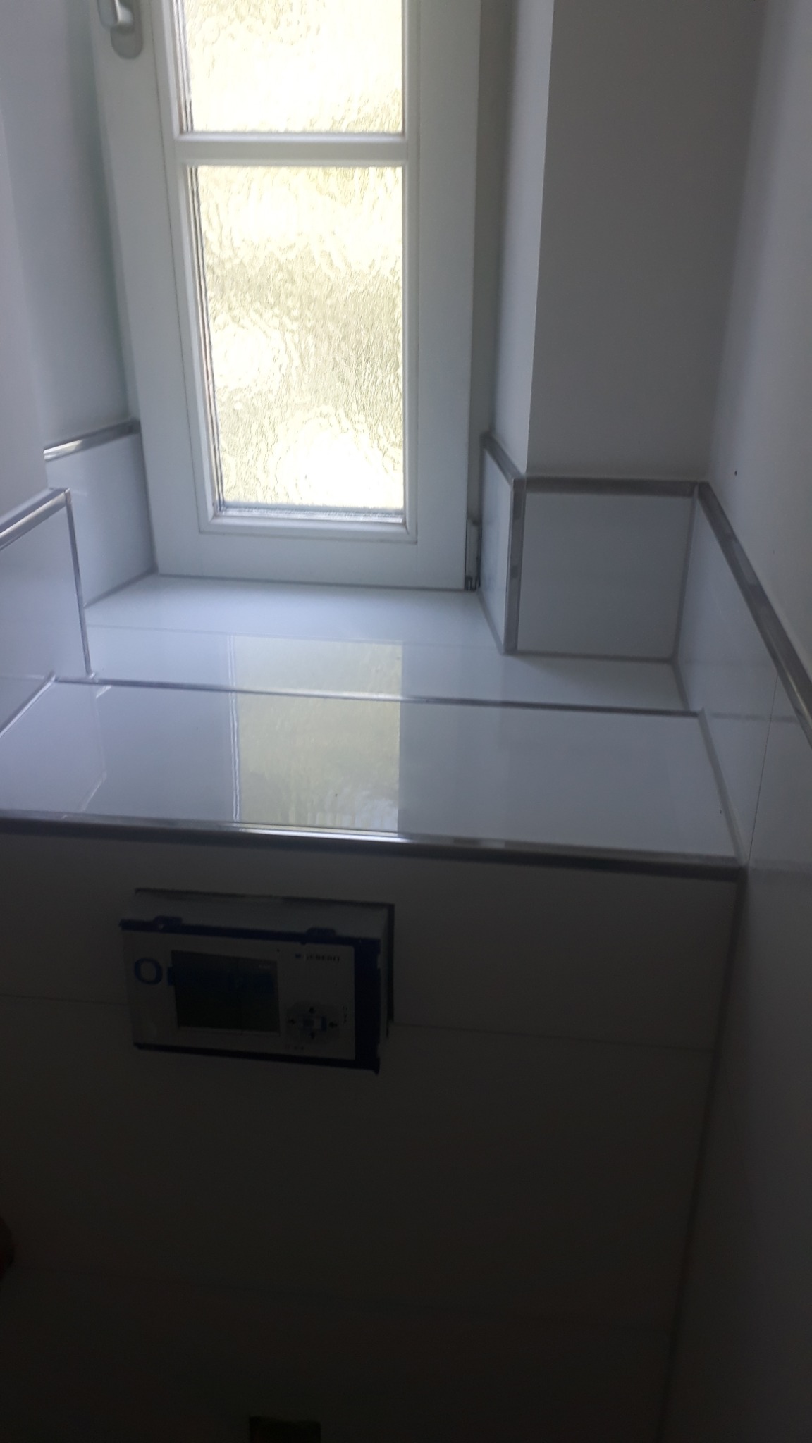 Biała, wykafelkowana półka pod oknem w łazience z zamontowanym sterownikiem do spłuczki, widok od dołu.