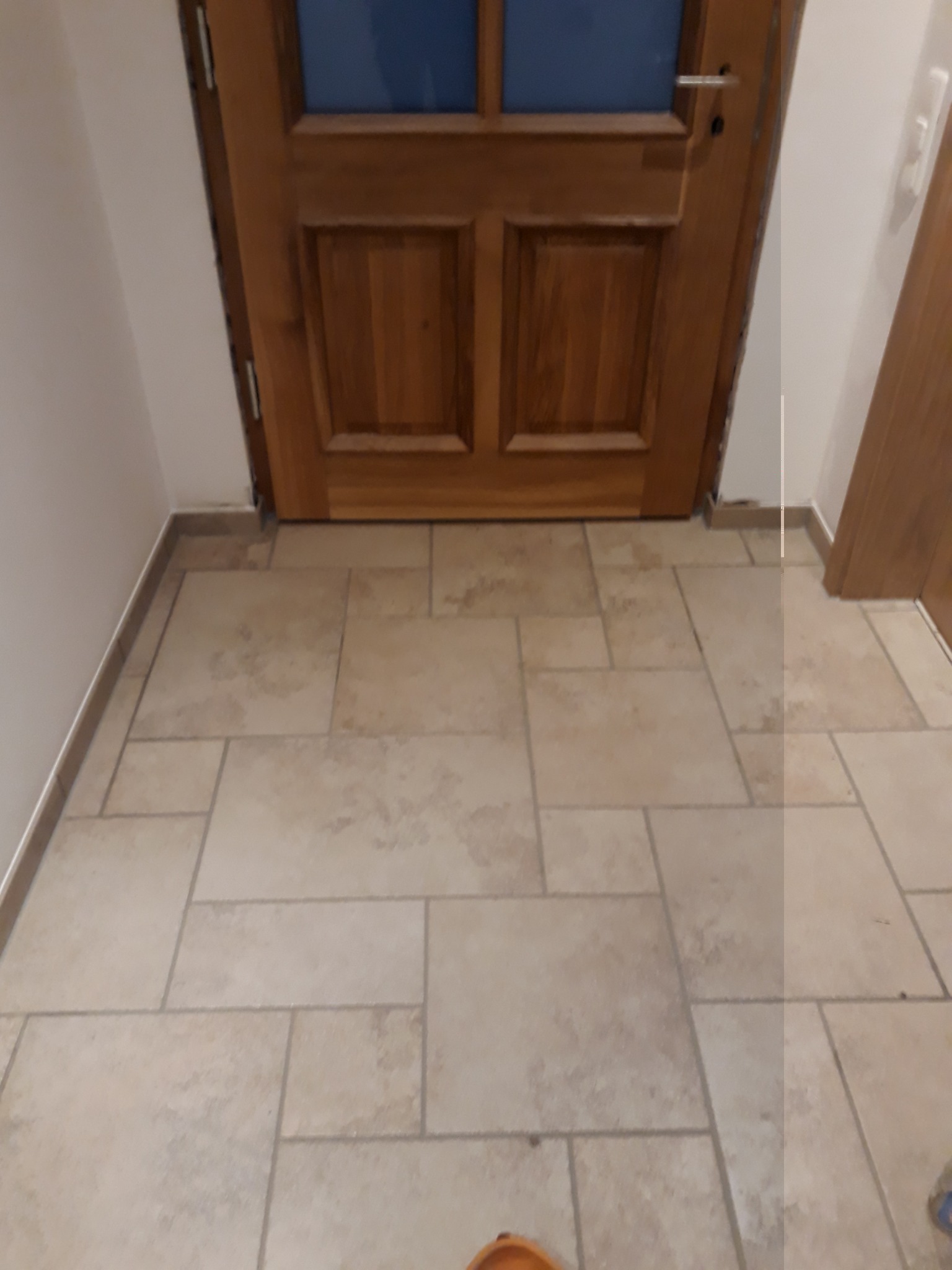 Wyłożona beżowymi płytkami podłoga w przedpokoju, widoczne drzwi z drewnianą ramą i szybami, oraz fragment ściany.