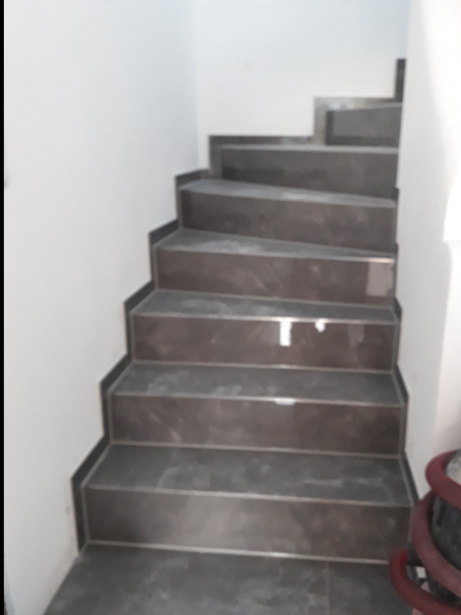 Wykończone schody wewnętrzne z ciemnoszarego marmuru z subtelnym wzorem, widoczne z dołu, z fragmentem ściany pomalowanej na biało oraz elementem węża odkurzacza w prawym dolnym rogu.