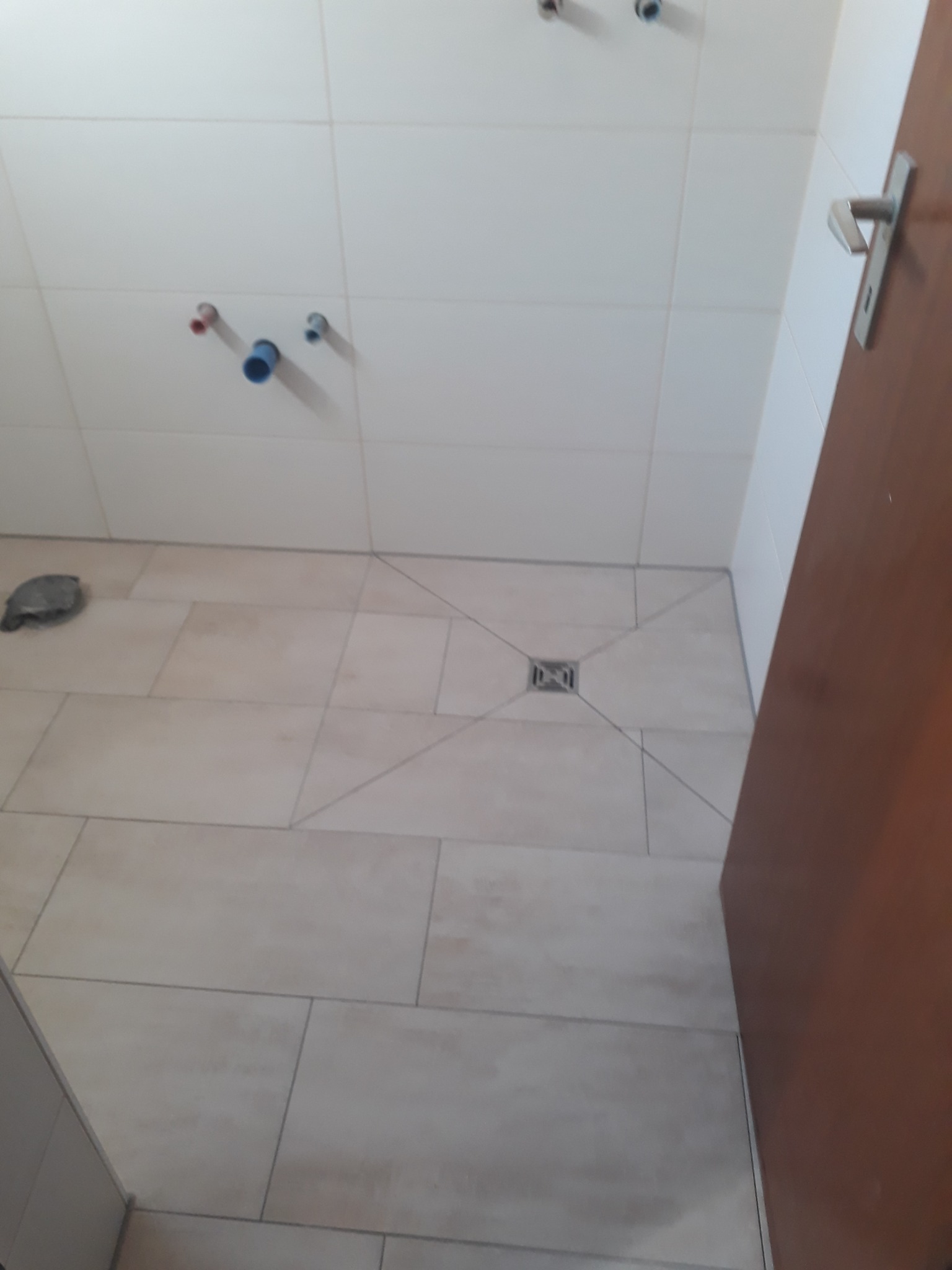 Łazienka w trakcie wykańczania: jasne płytki podłogowe i ścienne, widoczne rury instalacji wodnej i kanalizacyjnej, odpływ liniowy w podłodze, fragment brązowych drzwi.