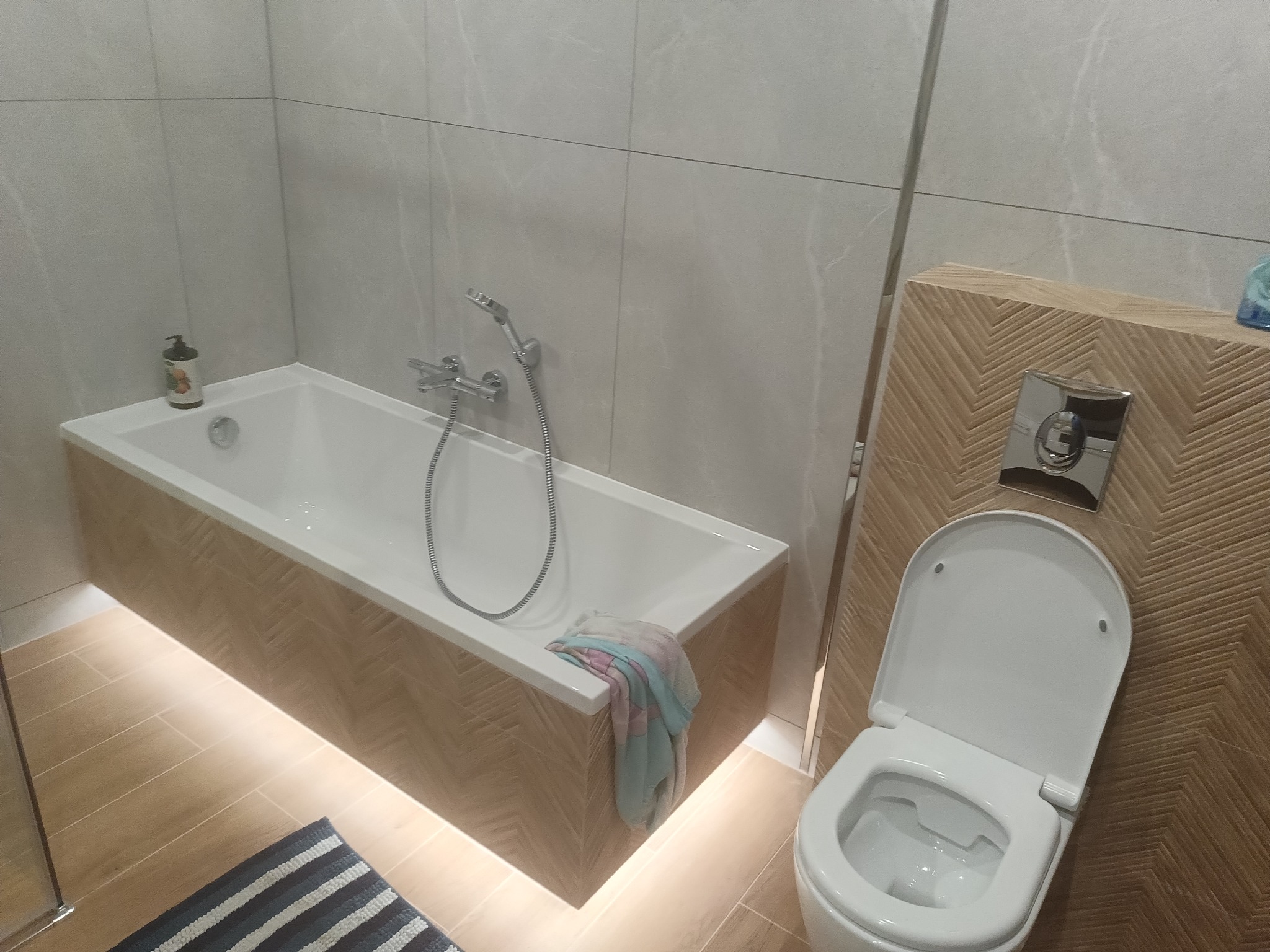 Nowoczesna łazienka z podświetlaną wanną obudowaną drewnopodobną płytką w jodełkę, wisząca toaleta z ukrytym stelażem i szarobiały dywanik w paski na podłodze z płytek imitujących drewno.