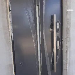 Nowoczesne, grafitowe drzwi wejściowe z długim, prostokątnym uchwytem, częściowo zabezpieczone folią ochronną, zamontowane w surowym otworze ściennym.