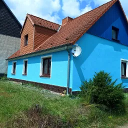 Dom z niebieską elewacją i brązowym dachem, widoczna antena satelitarna, obok rosnąca zieleń i częściowo widoczny biały samochód dostawczy.