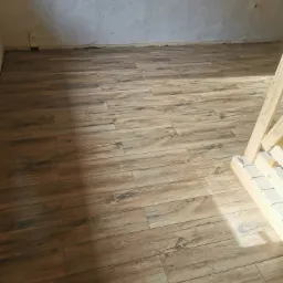 Ułożona podłoga z płytek imitujących drewno w jasnym odcieniu, widoczne ślady po fugowaniu na ścianie, fragment drewnianej konstrukcji po prawej stronie.