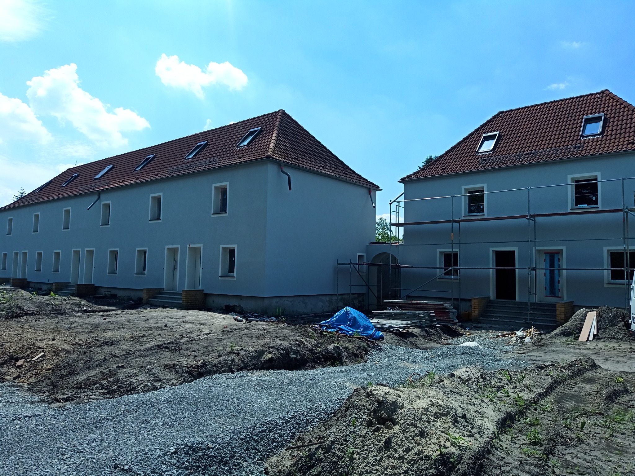 Dwa budynki w trakcie prac elewacyjnych z widocznymi rusztowaniami, niezagospodarowany teren wokół, błękitne niebo z chmurami w tle.