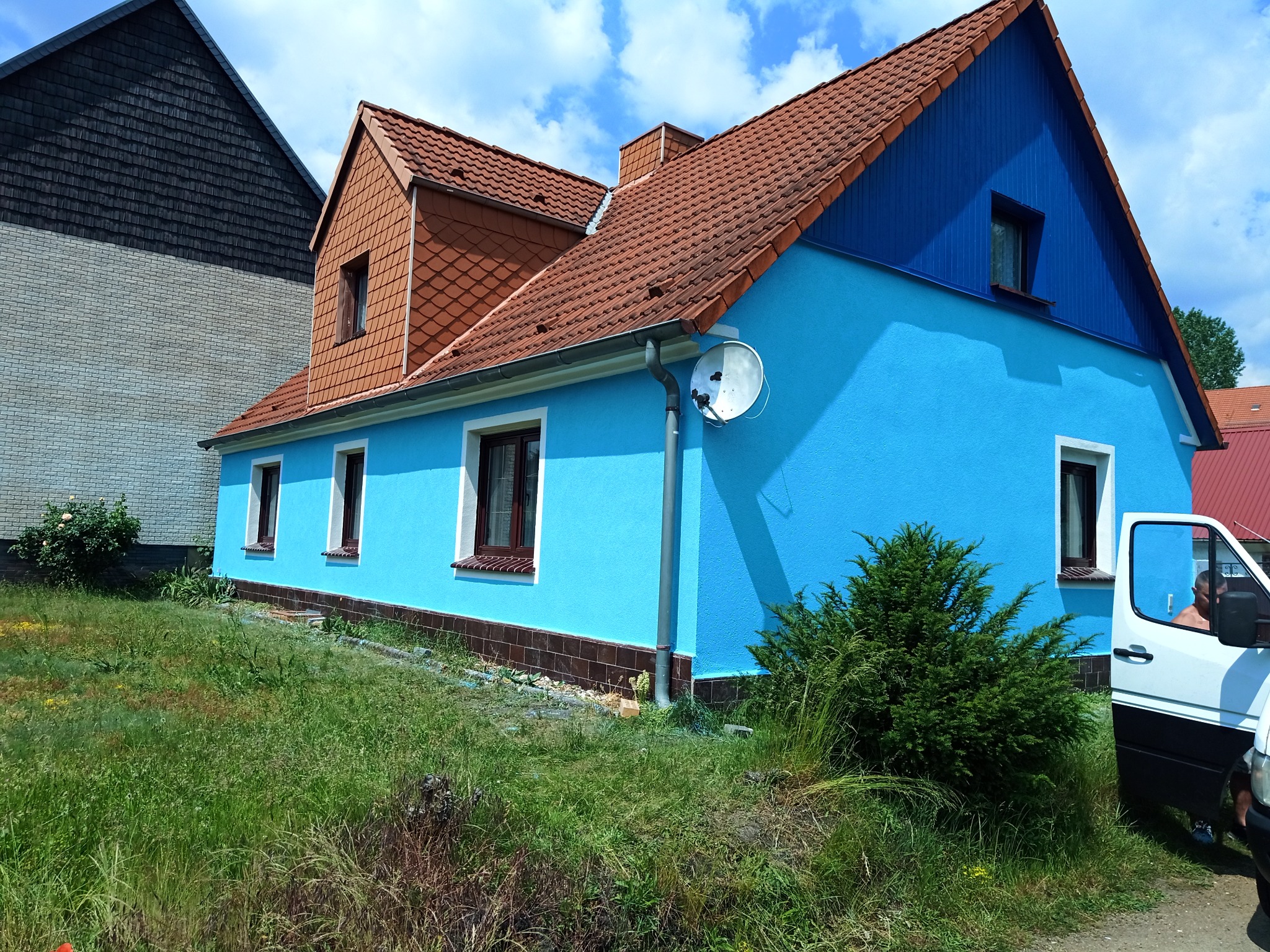 Dom z niebieską elewacją i brązowym dachem, widoczna antena satelitarna, obok rosnąca zieleń i częściowo widoczny biały samochód dostawczy.