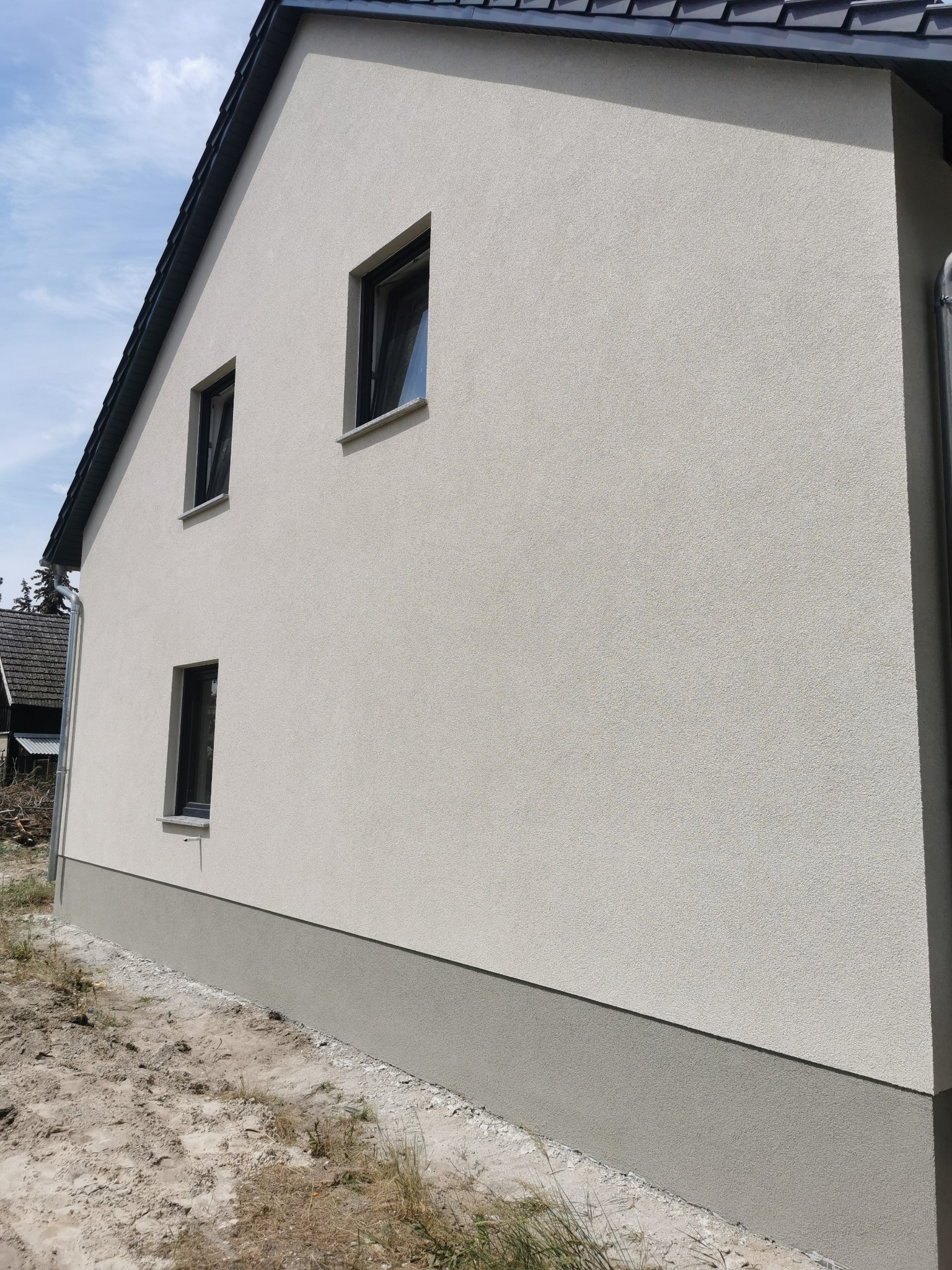 Ściana boczna domu jednorodzinnego z nową, jasną elewacją o fakturze baranka, z trzema oknami w czarnych ramach i szarą podmurówką, widok z zewnątrz na tle błękitnego nieba.