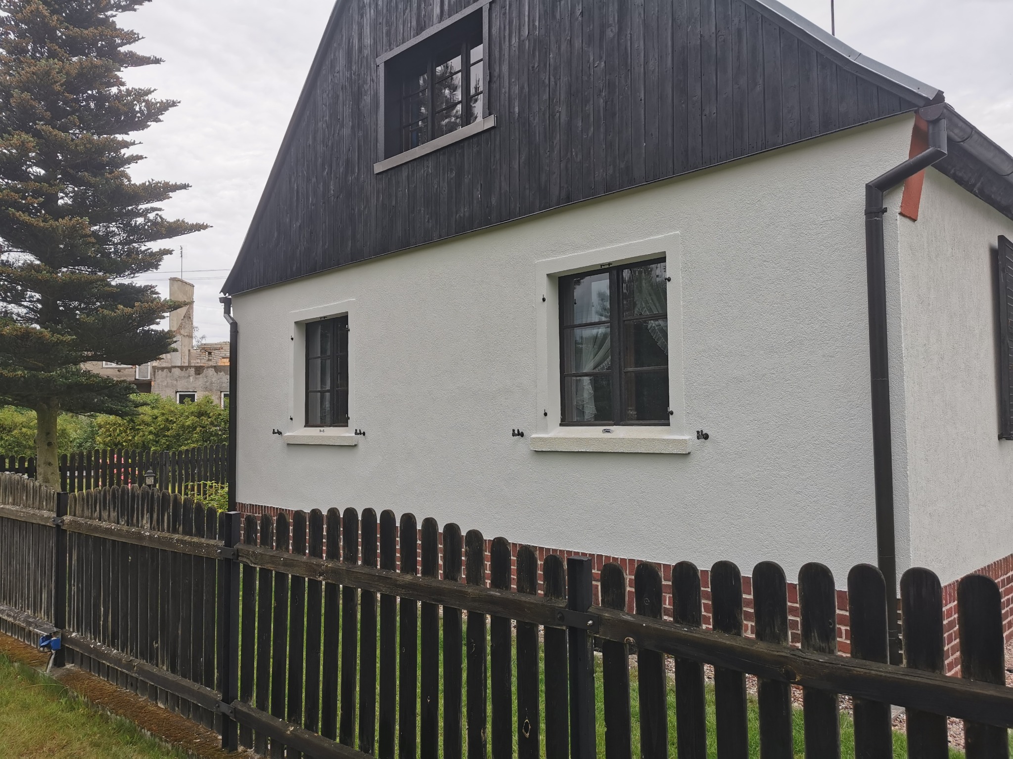 Biała elewacja domu z czarnymi drewnianymi elementami, widok zza ciemnego drewnianego płotu.
