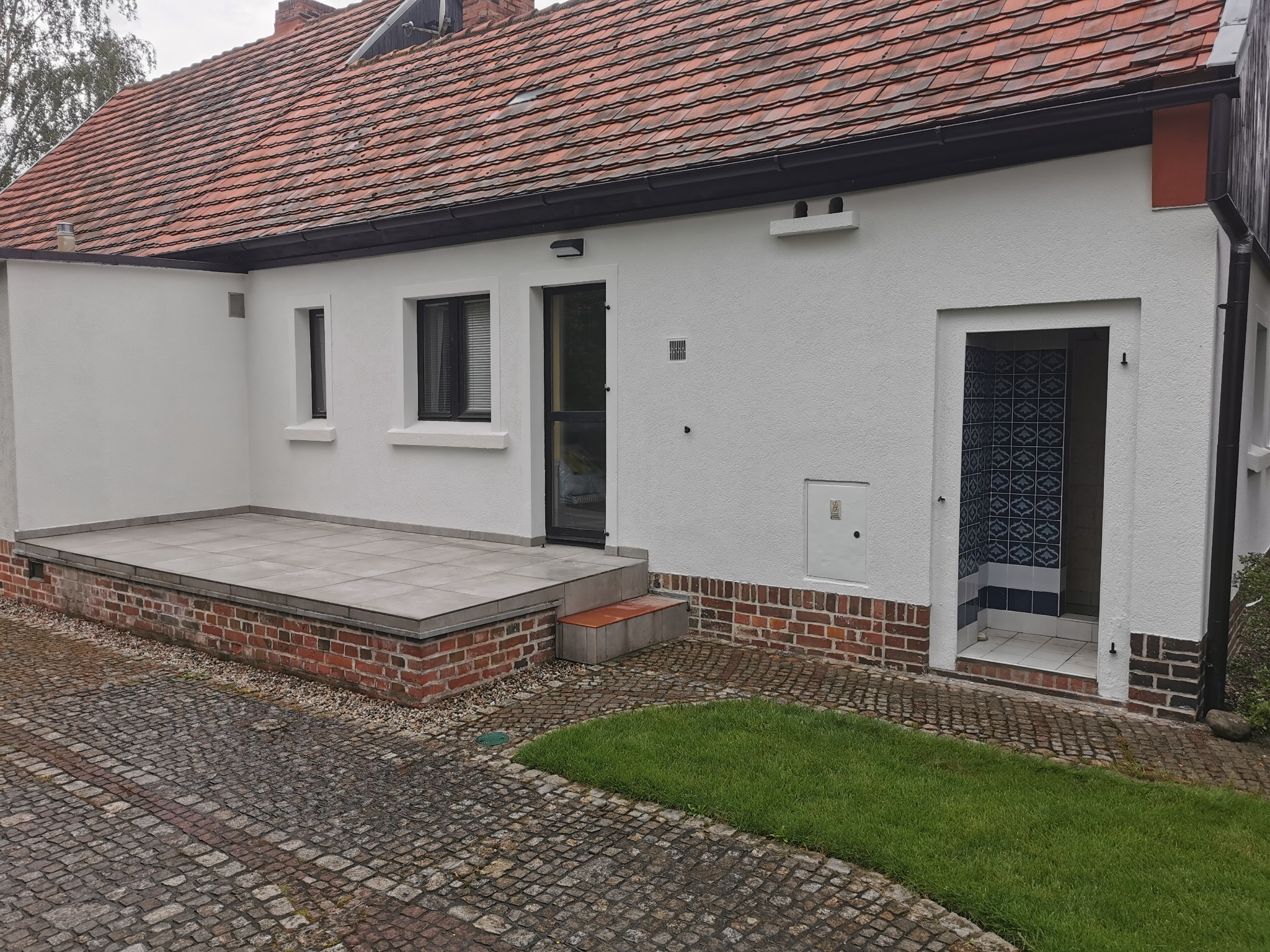 Biała elewacja budynku z czerwonym dachem, widoczny taras z szarymi płytkami, ceglana podmurówka, kostka brukowa na podwórku i fragment trawnika.