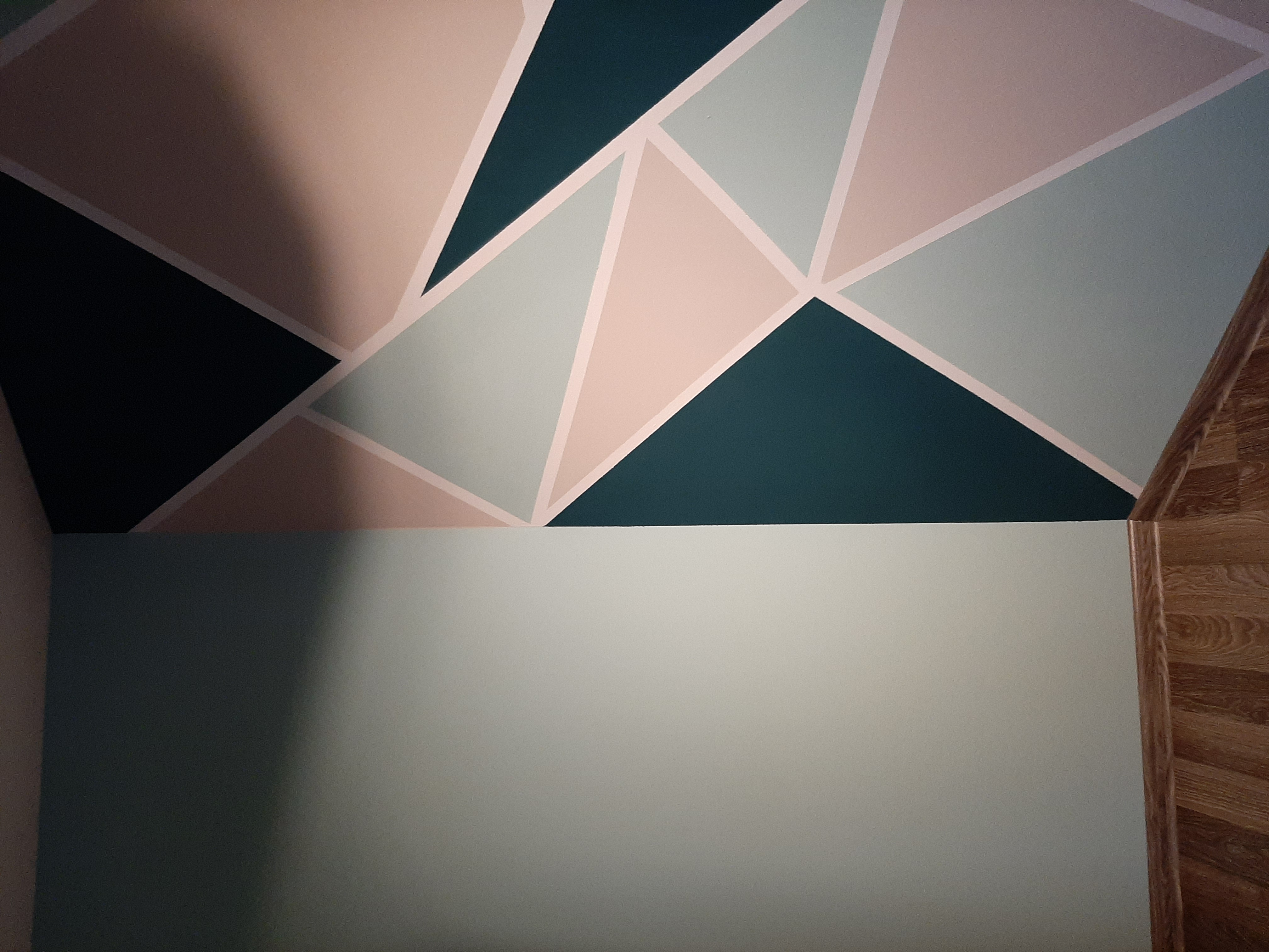 Ściana w pokoju z geometrycznym wzorem z trójkątów w odcieniach zieleni, beżu i szarości, oddzielona od jednolitej, jasnej dolnej części ściany. Widoczne wykończenie podłogi przy ścianie.