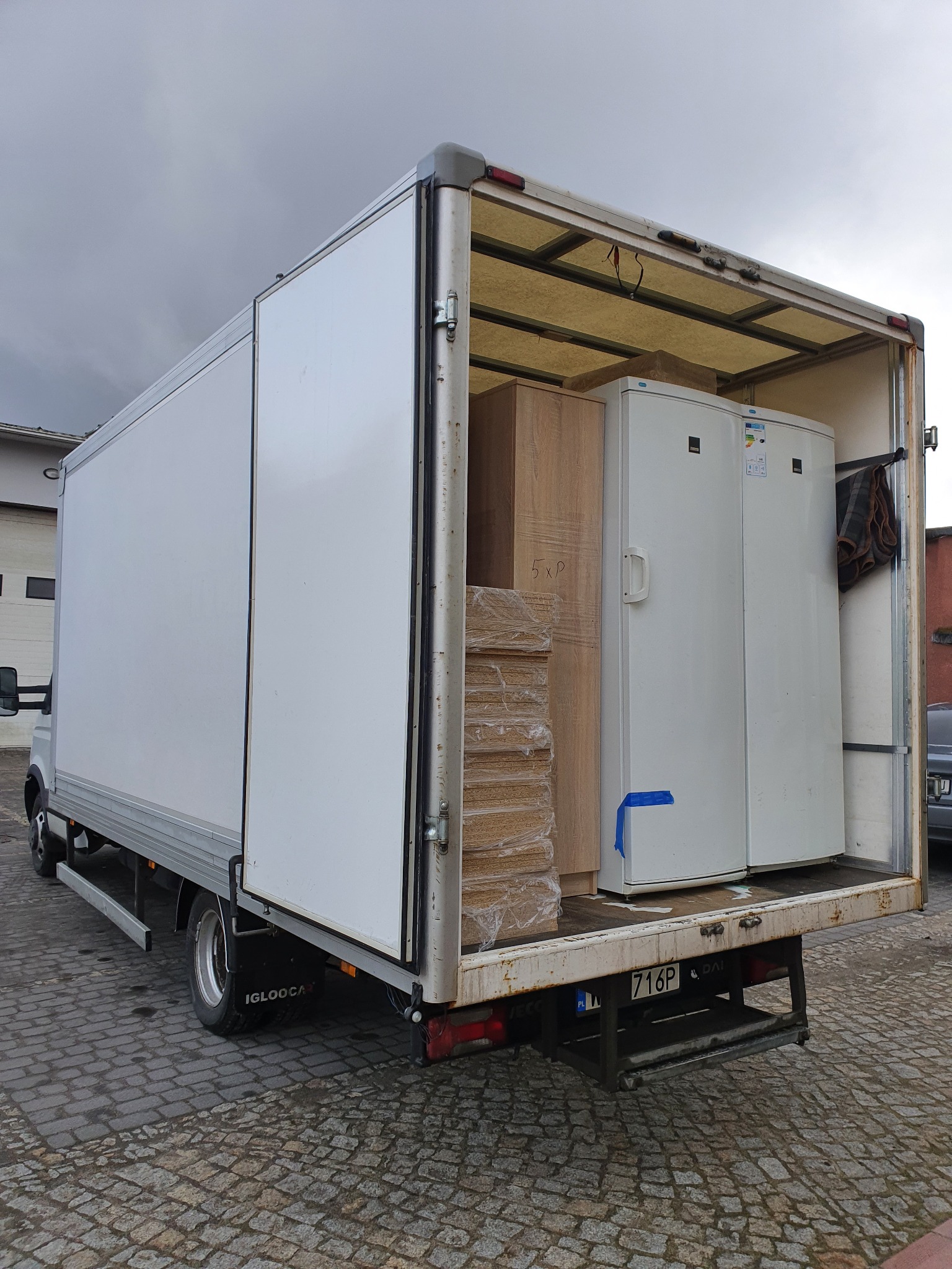 Otwarty tył białego samochodu dostawczego w Warszawie, załadowanego spakowanymi meblami i podwójną lodówką z naklejkami energetycznymi. Widoczny numer rejestracyjny pojazdu.