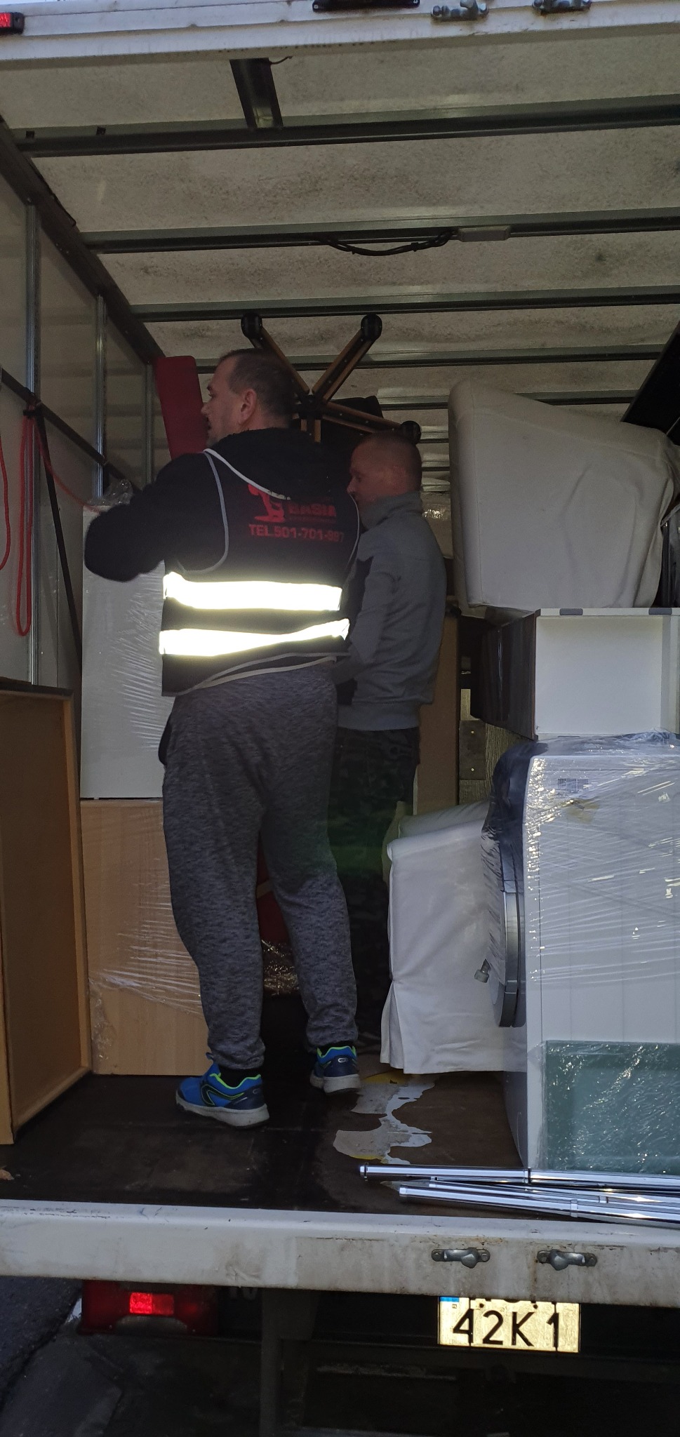 Dwóch mężczyzn w odblaskowych kamizelkach umieszcza meble owinięte folią wewnątrz furgonetki transportowej podczas przeprowadzki, widoczny numer rejestracyjny 42K1 pojazdu.