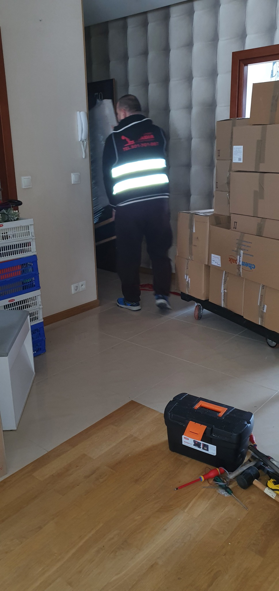 Pracownik w kamizelce odblaskowej wnosi zapakowany przedmiot do mieszkania, obok sterta kartonów na wózku, skrzynki i narzędzia na podłodze.