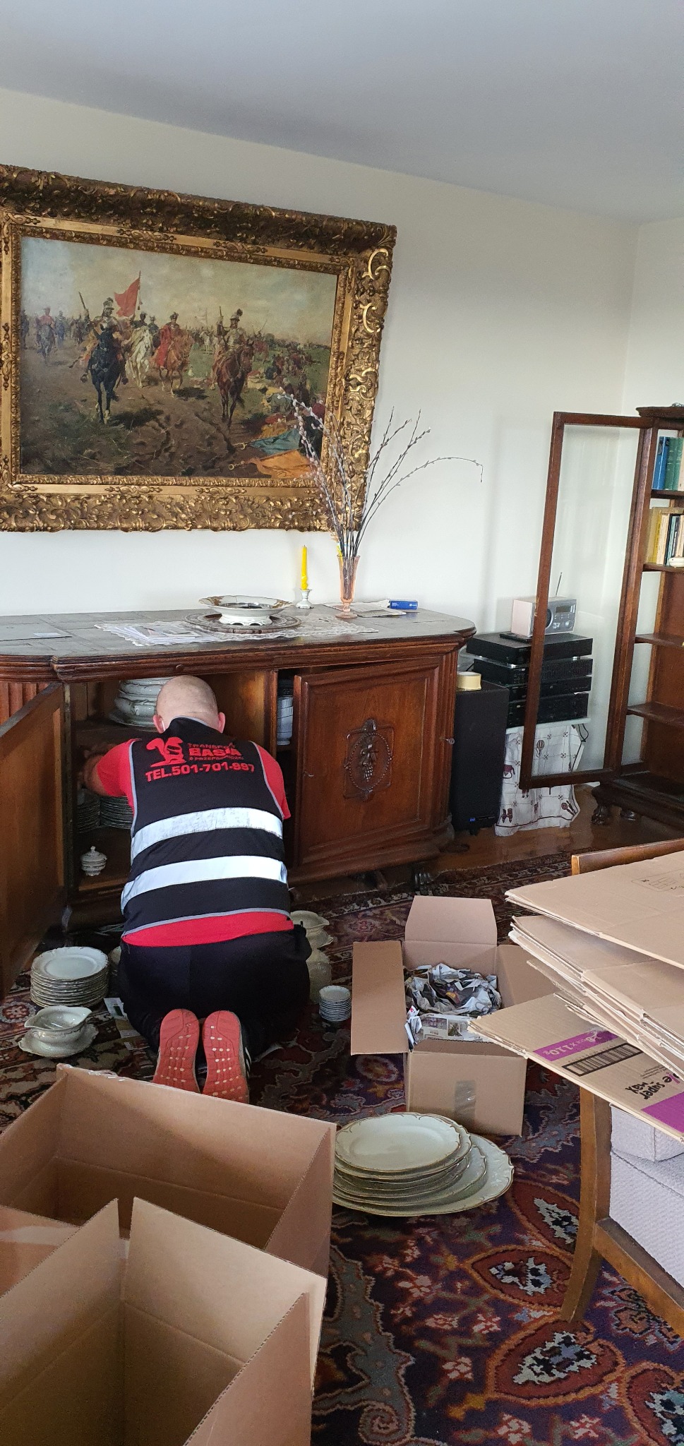 Pracownik w odblaskowej kamizelce klęczy i ostrożnie wyjmuje talerze z kredensu, przygotowując je do spakowania w kartonowe pudła podczas przeprowadzki. W tle widoczny obraz w złotej ramie...