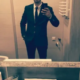 Mężczyzna w garniturze robiący selfie w łazience, widoczne odbicie w lustrze nad umywalką z dozownikiem mydła i ręcznikami papierowymi.