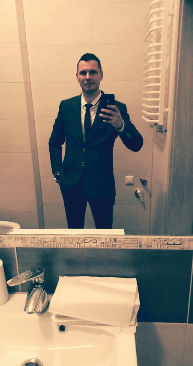 Mężczyzna w garniturze robiący selfie w łazience, widoczne odbicie w lustrze nad umywalką z dozownikiem mydła i ręcznikami papierowymi.