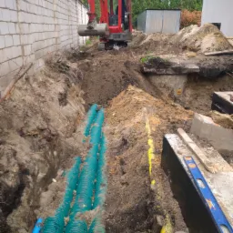 Czerwona mini koparka na placu budowy, wykopane rowy z ułożonymi niebieskimi i żółtymi rurami karbowanymi, w tle ściana z bloczków i metalowy garaż.