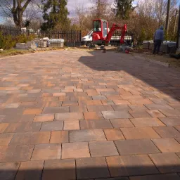 Ułożona kostka brukowa w odcieniach brązu i czerwieni, z widocznym mini-koparką Yanmar i stosem kostki w tle, częściowo zacieniona