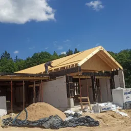 Budowa domu jednorodzinnego: murarz układa płyty OSB na drewnianej konstrukcji dachu, widoczne bloczki Solbet, piasek i niebo z chmurami.