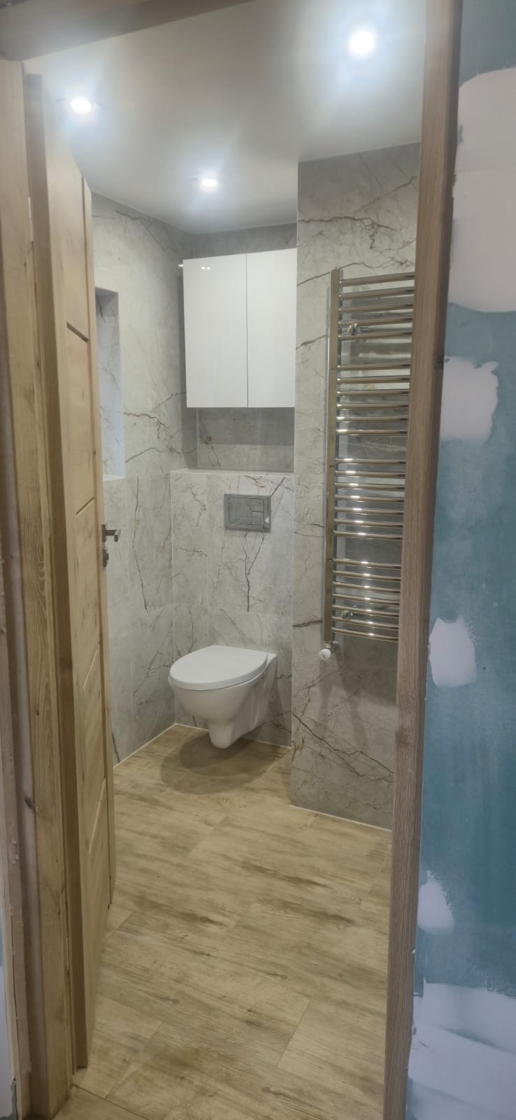 Wykończona łazienka z marmurowymi płytkami, podwieszaną toaletą i grzejnikiem drabinkowym. Widok zza drewnianej ościeżnicy, ściana po prawej w trakcie malowania.