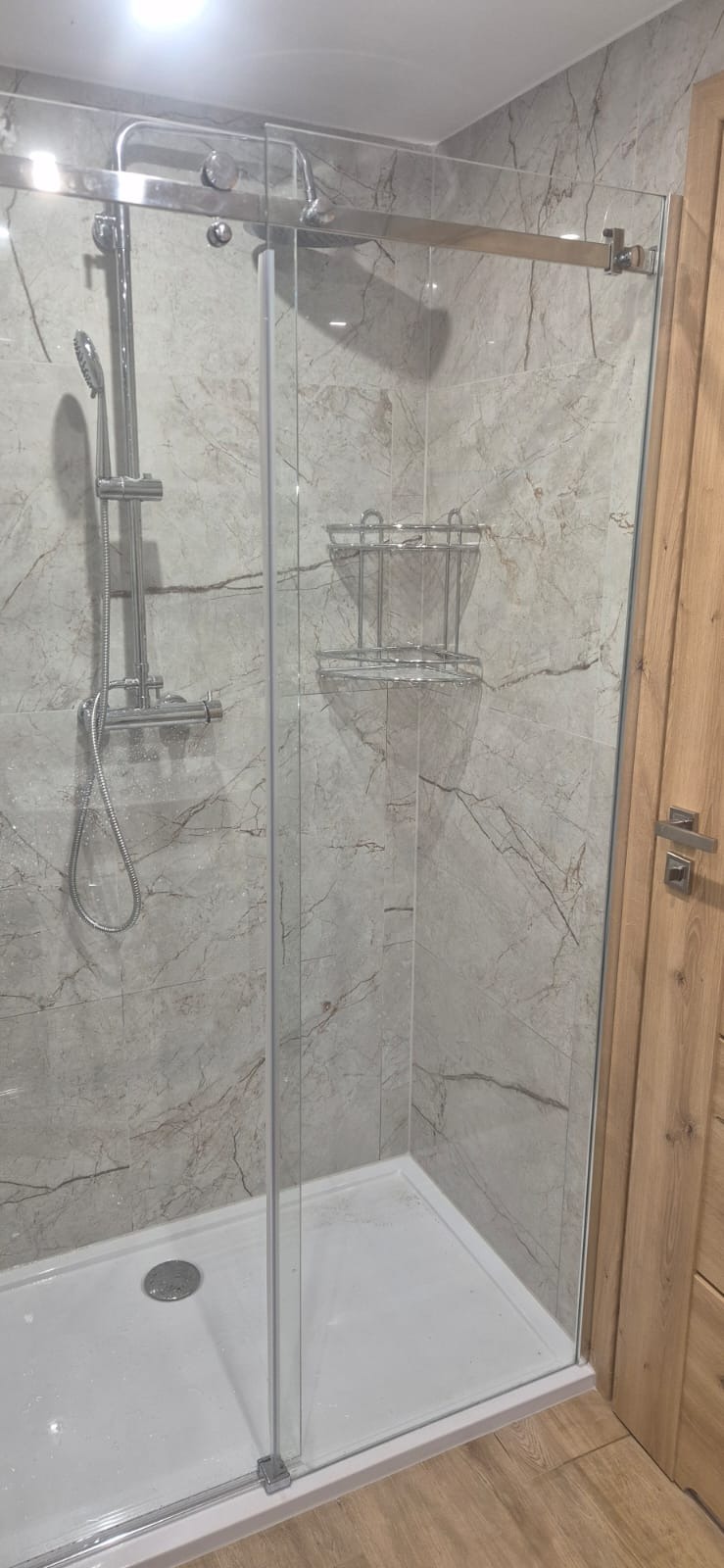 Nowoczesna kabina prysznicowa z przesuwnymi drzwiami, wykończona płytkami imitującymi marmur i drewnianymi elementami. Widoczna armatura i półka na kosmetyki.