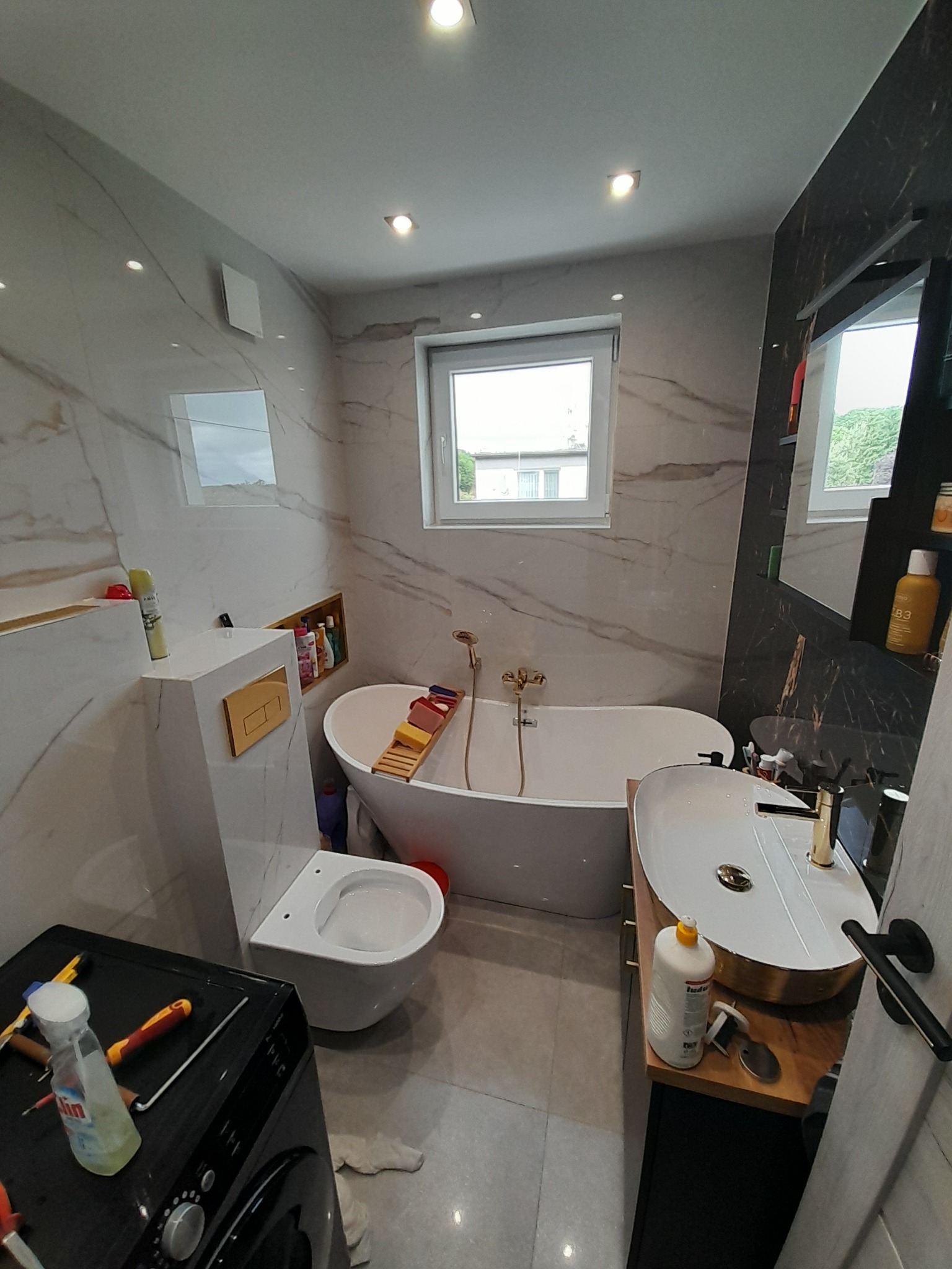 Elegancka łazienka z wanną wolnostojącą, umywalką nablatową ze złotymi akcentami i marmurowymi płytkami. Widoczne narzędzia sugerują trwające prace wykończeniowe.