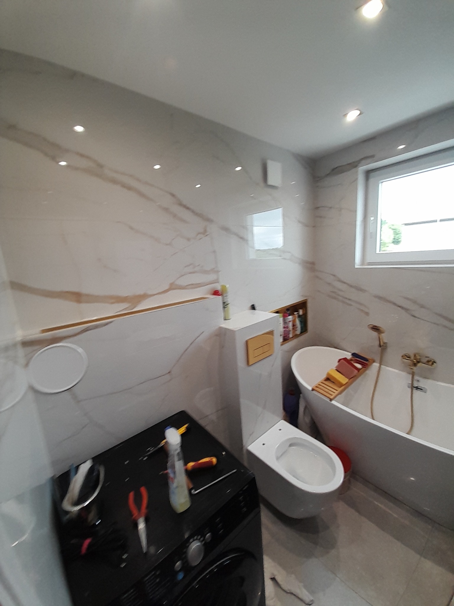 Elegancka łazienka z marmurowymi płytkami, wolnostojącą wanną i podwieszaną toaletą. Na pralce widoczne narzędzia, sugerujące trwające prace wykończeniowe.