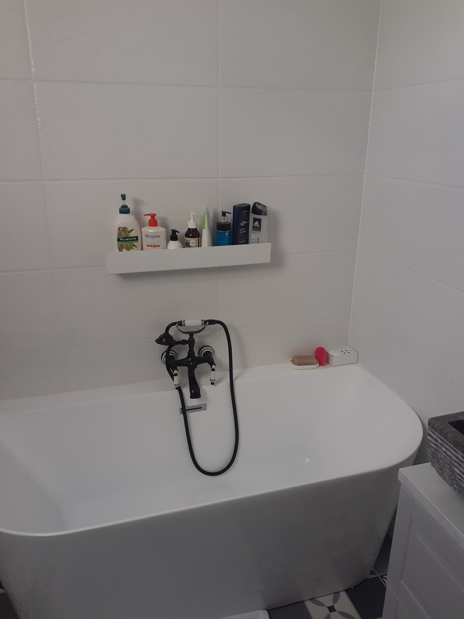 Biała, owalna wanna z czarną armaturą retro, białe płytki na ścianie i półka z kosmetykami w nowoczesnej łazience.