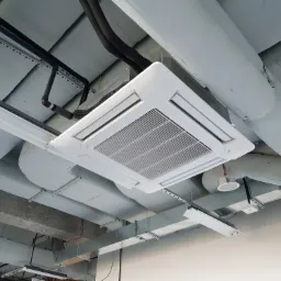 Kasetonowa jednostka klimatyzacyjna zamontowana pod sufitem w biurze, widoczne rury instalacyjne i oświetlenie liniowe.