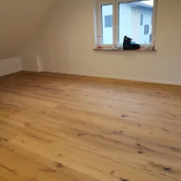 Ułożona podłoga z paneli drewnopodobnych w nowym, jasnym pokoju na poddaszu z oknem, widoczne gniazdka elektryczne i elementy instalacji.