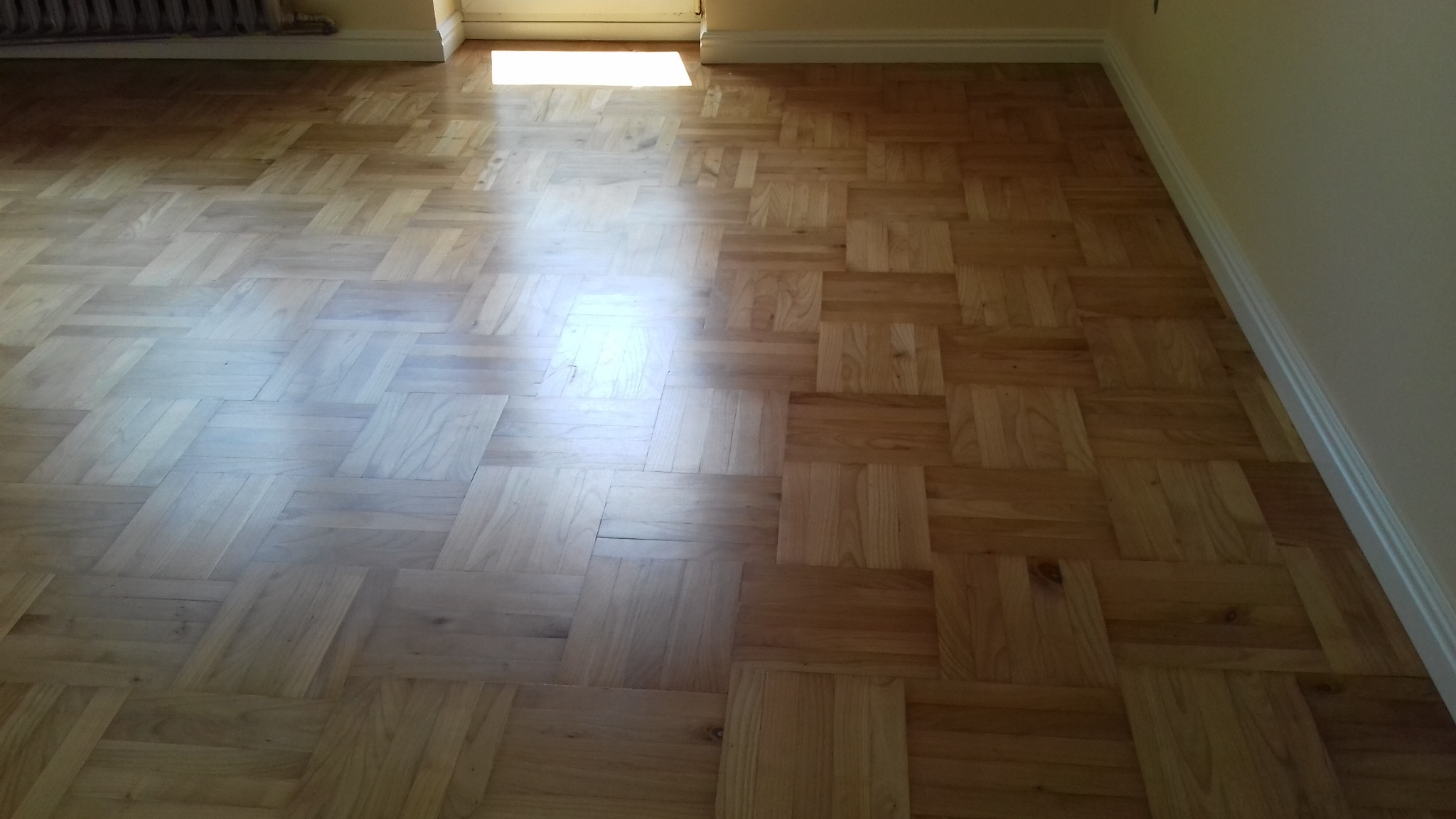 Jasny parkiet ułożony we wzór kwadratów, wykończony białą listwą przypodłogową, oświetlony naturalnym światłem wpadającym przez drzwi.