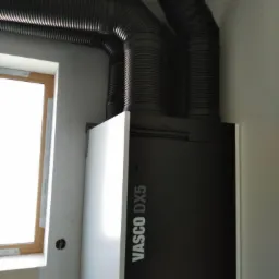 Czarna jednostka rekuperacyjna Vasco DX5 z podłączonymi karbowanymi przewodami wentylacyjnymi, widoczna obok okna w surowym wnętrzu.