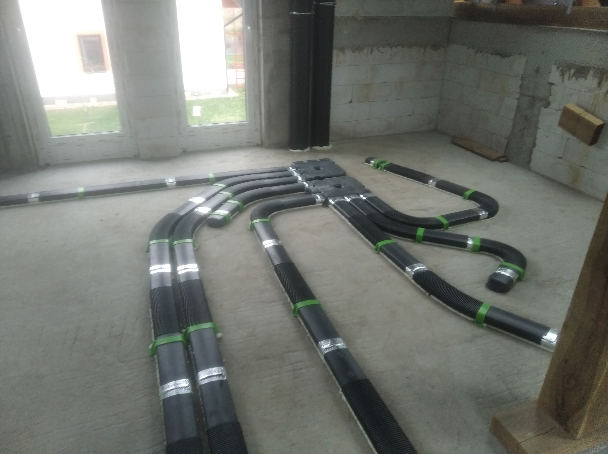 Rozłożone czarne rury wentylacyjne z zielonymi opaskami i srebrną taśmą na podłodze w surowym pomieszczeniu z oknem balkonowym.