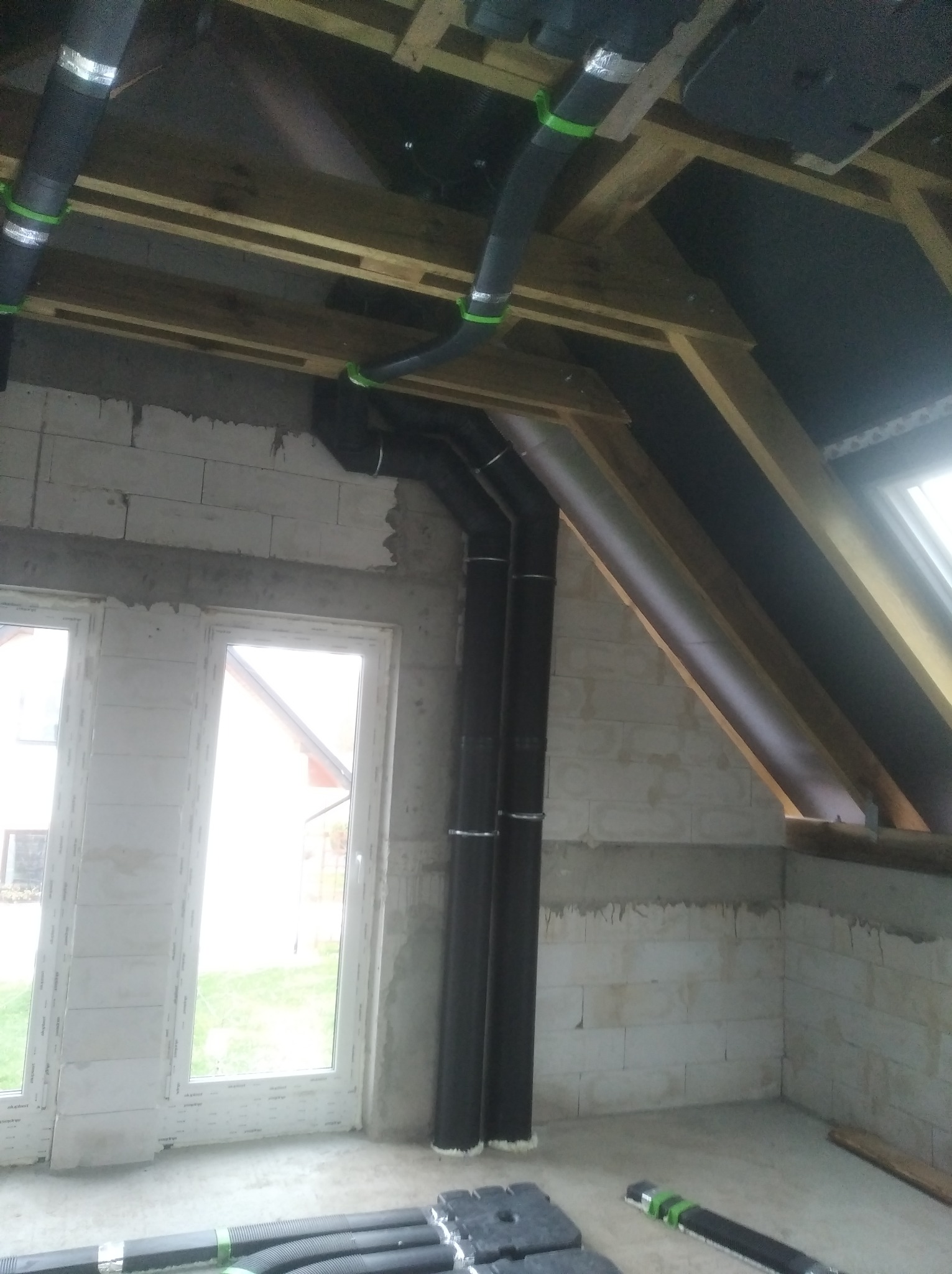 Wnętrze w trakcie montażu instalacji wentylacyjnej z widocznymi rurami wentylacyjnymi pod sufitem, mocowanymi do drewnianej konstrukcji, z elementami izolacji i fragmentem okna w tle.