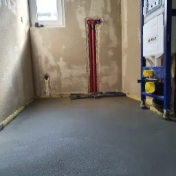 Świeżo wylana, szara posadzka w surowym wnętrzu łazienki z widocznymi instalacjami hydraulicznymi w ścianie i stelażem podtynkowym Geberit.