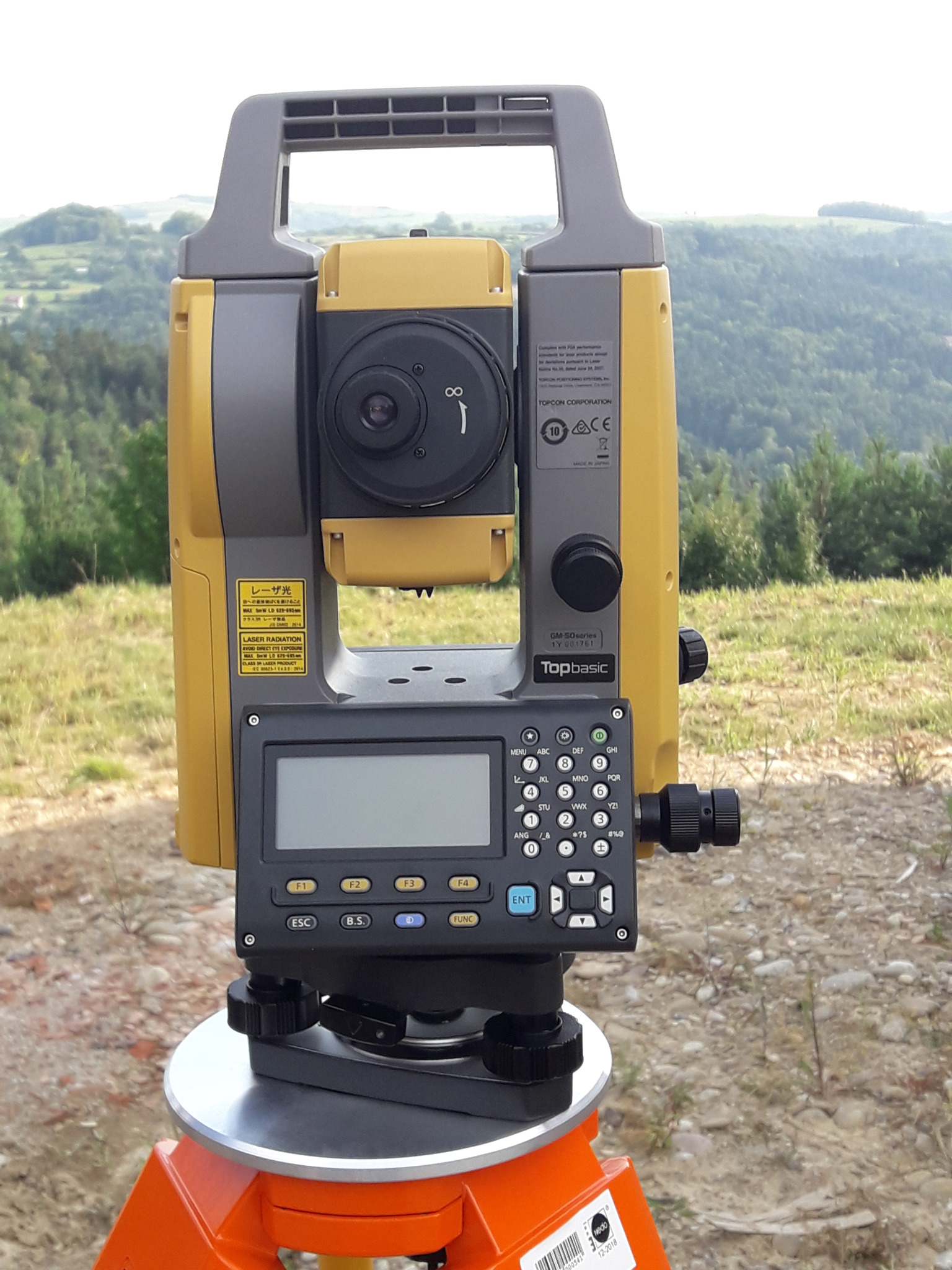Żółto-szary tachimetr Topbasic na pomarańczowym statywie, ustawiony na zewnątrz z widokiem na zalesione wzgórza w tle. Urządzenie gotowe do pomiarów.