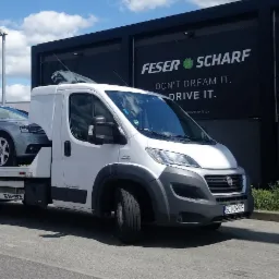 Biała laweta Fiat Ducato z srebrnym kombi Audi A4 na platformie, zaparkowana przed salonem Land Rover i Feser Scharf w słoneczny dzień.