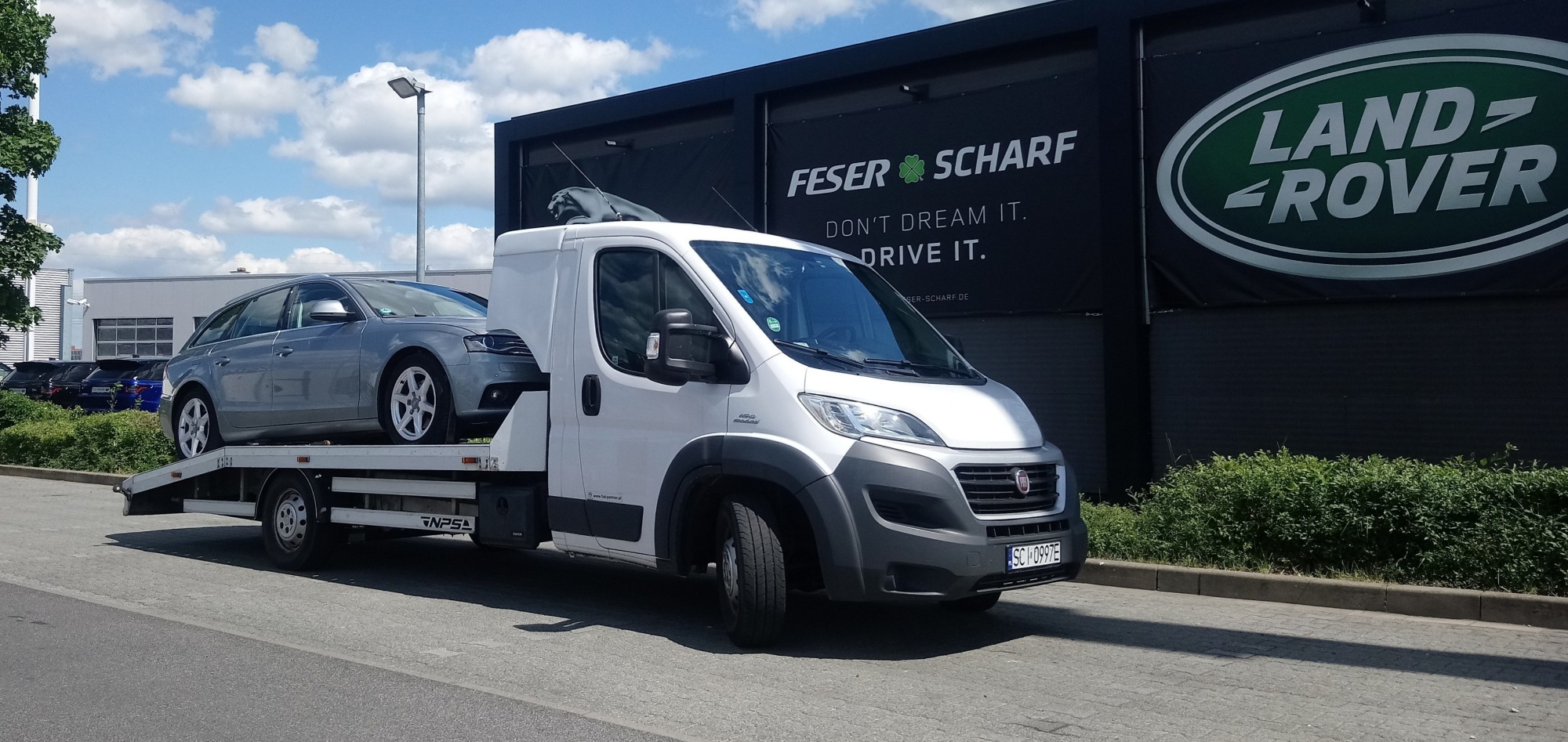 Biała laweta Fiat Ducato z srebrnym kombi Audi A4 na platformie, zaparkowana przed salonem Land Rover i Feser Scharf w słoneczny dzień.