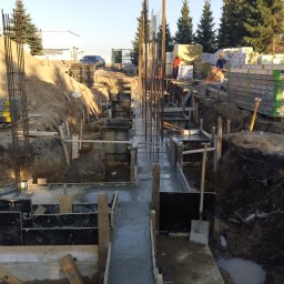 Baujp Budownictwo - Wylane betonowe fundamenty z wystającymi prętami zbrojeniowymi, szalunki z desek, szczotka do czyszczenia na betonie, widoczni pracownicy w oddali oraz stosy materiałów budowlanych w tle.