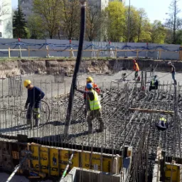 Wylewanie betonu na zbrojenie fundamentów budynku, widoczne szalunki Doka, pracownicy w kaskach i kamizelkach odblaskowych, na drugim planie budynek mieszkalny i drzewa.