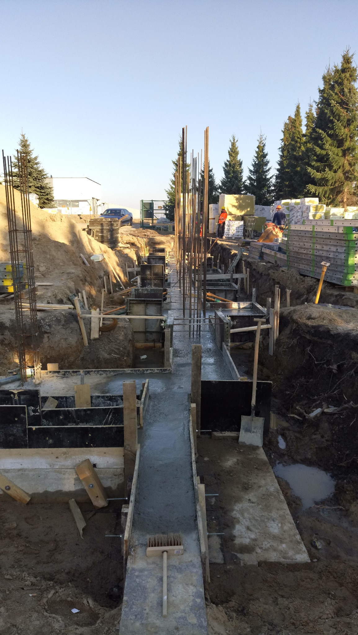 Wylane betonowe fundamenty z wystającymi prętami zbrojeniowymi, szalunki z desek, szczotka do czyszczenia na betonie, widoczni pracownicy w oddali oraz stosy materiałów budowlanych w tle.