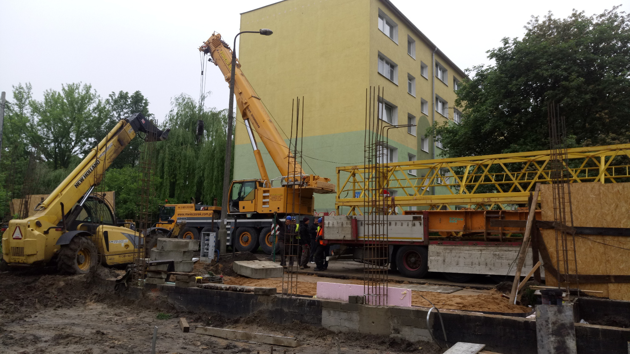 Prace ziemne pod fundamenty w Łodzi: widok na plac budowy z ciężkim sprzętem, w tym dźwigiem i podnośnikiem teleskopowym, oraz grupą pracowników w pobliżu wykopu.