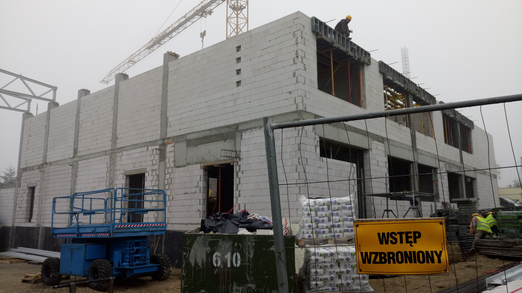 Wznoszenie dwukondygnacyjnego budynku z bloczków, widoczny żuraw budowlany w tle i pracownik na górnej kondygnacji, tablica ostrzegawcza z napisem 'Wstęp wzbroniony' na pierwszym planie.