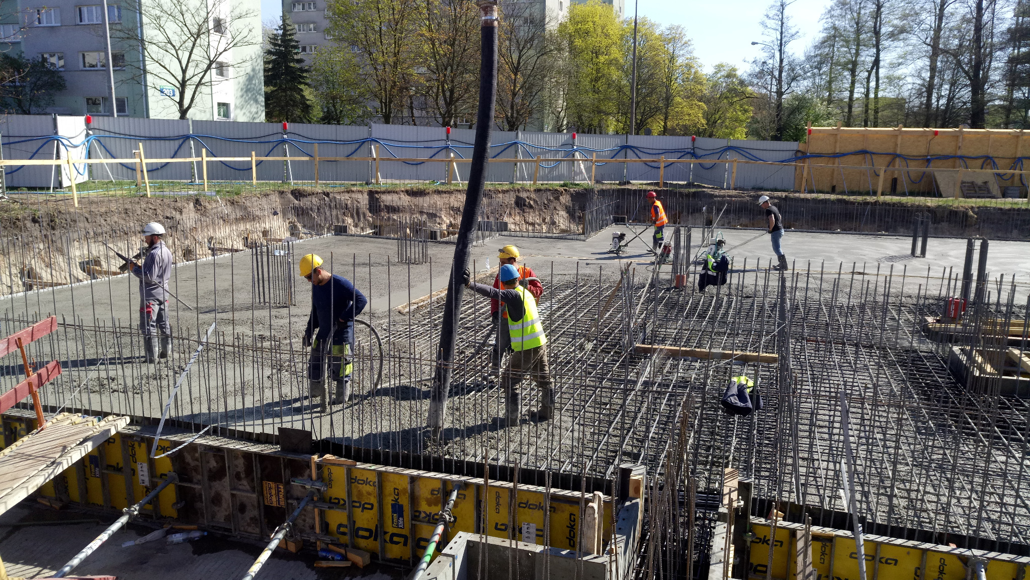 Wylewanie betonu na zbrojenie fundamentów budynku, widoczne szalunki Doka, pracownicy w kaskach i kamizelkach odblaskowych, na drugim planie budynek mieszkalny i drzewa.