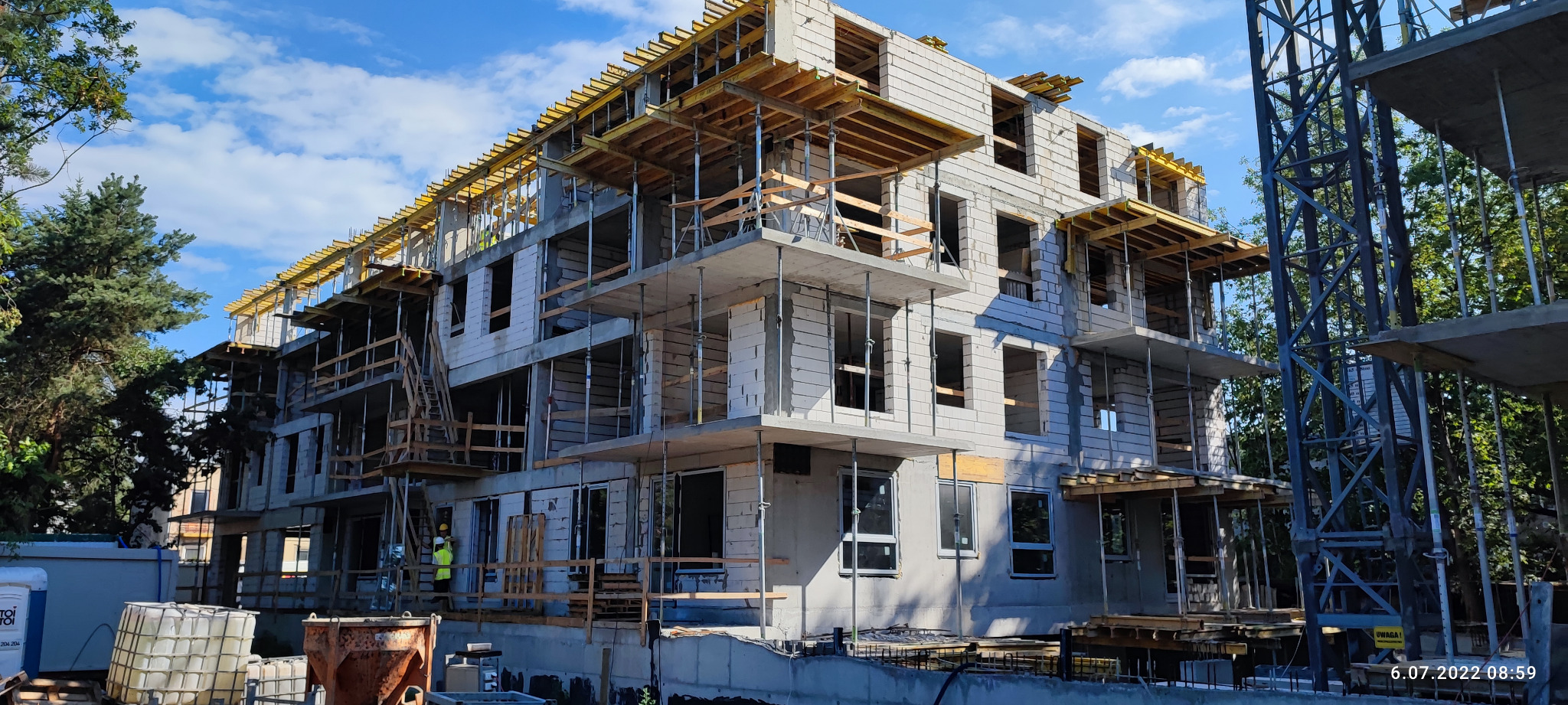 Wielopiętrowy budynek mieszkalny w stanie surowym, w trakcie budowy, z widocznymi elementami szalunków i rusztowań, wykonany z bloczków betonu komórkowego, z pracownikami w żółtych kamizelkach...