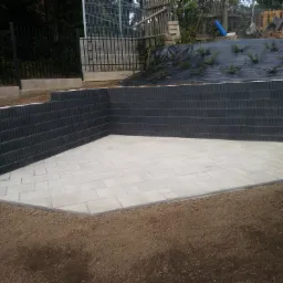 Kąt ujęcia na wykończony taras z jasnej kostki brukowej, otoczony niskim murem z ciemnej kostki, z widocznym przygotowanym terenem pod dalsze nasadzenia roślin.