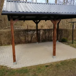 Zadaszone miejsce wypoczynku z metalowym dachem, drewnianymi słupami i brukowaną podłogą, otoczone krzewami i drzewami w tle.