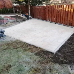 Przygotowany teren pod taras z ułożonymi płytami betonowymi, ubijarka wibracyjna obok, w tle drewniany płot i elementy przyszłego ogrodu.