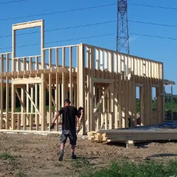 Wznoszenie szkieletu drewnianego domu jednorodzinnego na fundamencie, widok z tyłu na pracownika z kablem w dłoni, w tle słup wysokiego napięcia i błękitne niebo.